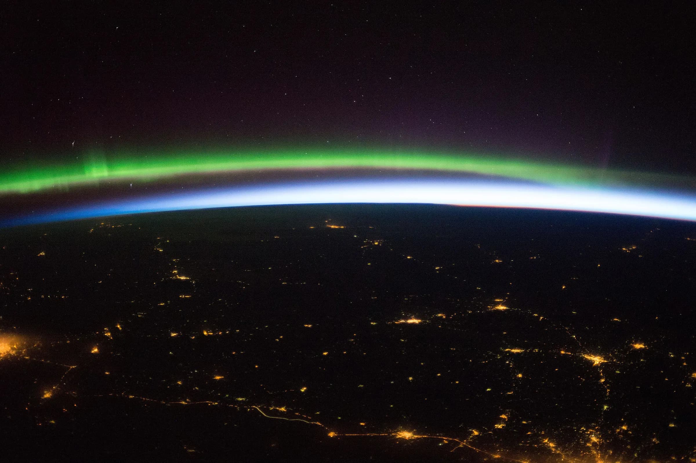 Northern Europe at night seen from the International Space Station, with city lights below and a green aurora on the horizon.