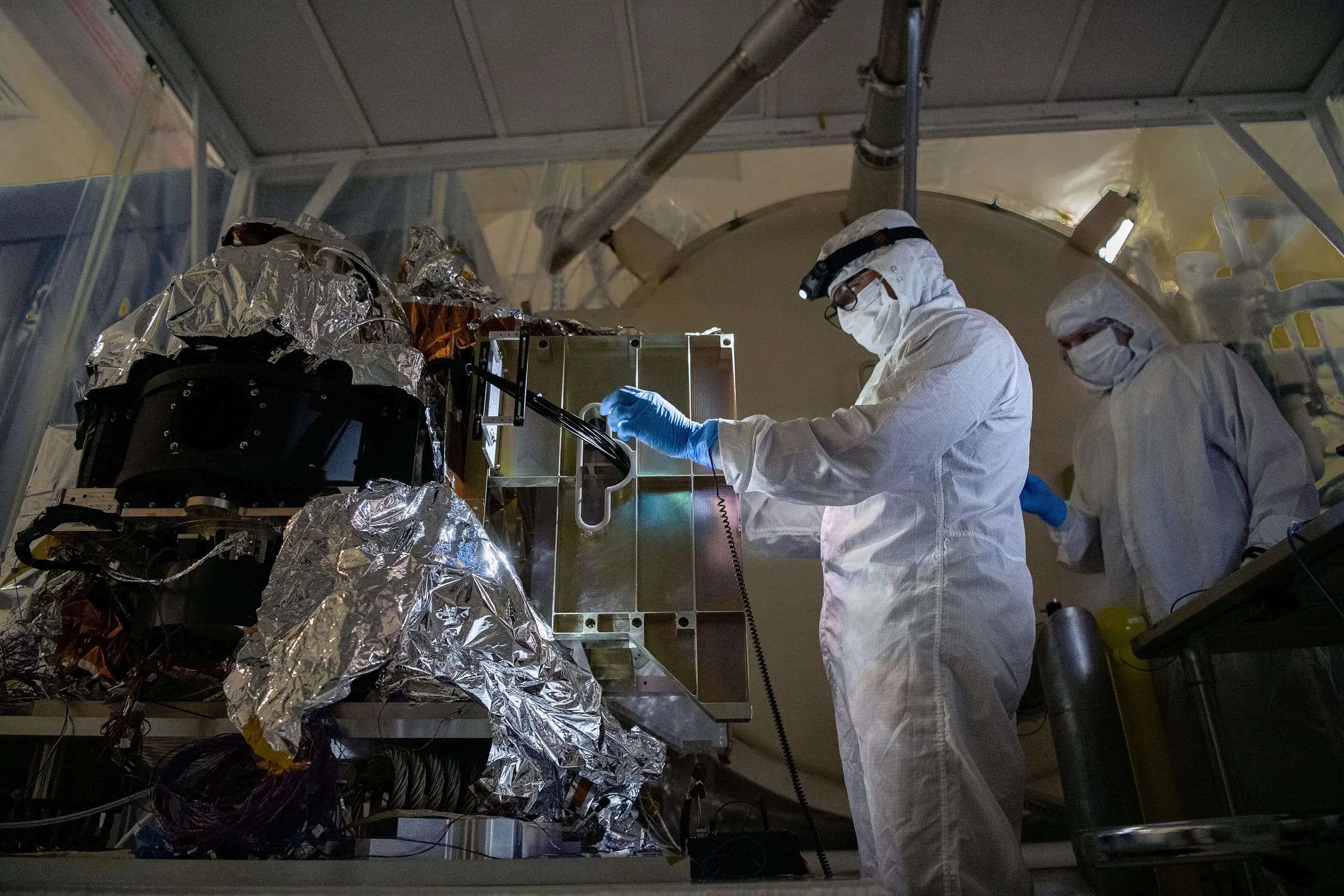 Engineers in cleanroom suits configuring optical fibers on a space instrument.